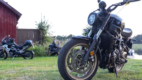 Im Vordergrund steht ein mit einem Bremsscheibenschloss gesichertes Motorrad. Im Hintergrund steht ein zweites Motorrad und zwei Menschen sitzen auf einer Bank. ©ABUS