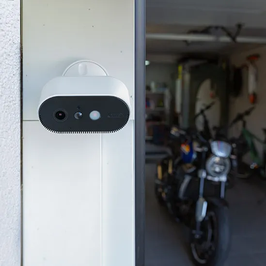 Eine weiße Überwachungskamera mit integriertem Licht ist an einer Garage angebracht, in der ein Motorrad parkt, und überwacht den Eingangsbereich. ©ABUS